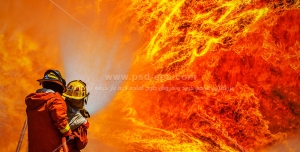 عکس با کیفیت آتش نشان در مقابل دیواری از شعله های مهیب آتش در حال خاموش کردن با شلنگ آتش نشانی و دو آتش نشان