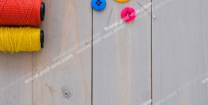 عکس با کیفیت قرقره های نخ های رنگی در کنار دکمه های رنگی و سوزن های چیده شده روی میز چوبی