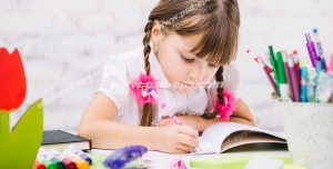 عکس با کیفیت تبلیغاتی دختر بچه زیبا با موهای بافته شده نشسته پشت میز در حال رنگ آمیزی و نقاشی