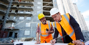 عکس با کیفیت تبلیغاتی مهندس ناظر در حال برسی نقشه در کنار یکی از کارگران ساختمان و ساختمان های بزرگ و مرتفع در پشت سرشان